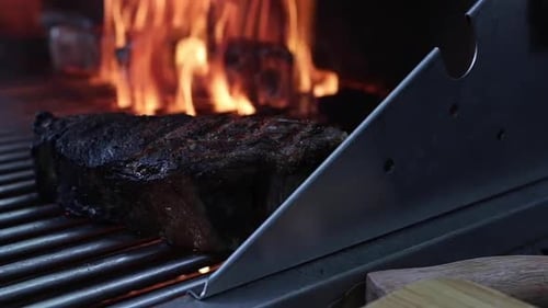 Flames rising on barbeque behind well done steak in foreground, close up shallow focus slow motion