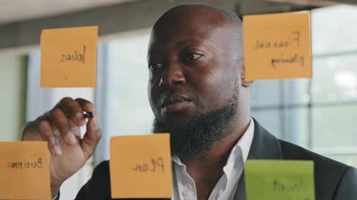 Afrikanischer Geschäftsmann steht im modernen Büro und schreibt Ideen auf transparente Wand mit Erinnerungsaufklebern