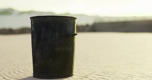 Black Trash Bin on Sandy Beach with Ocean Background During Sunset
