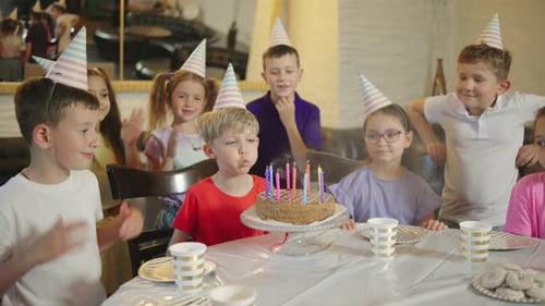 Um aniversariante com um boné festivo apagando as velas de um bolo de aniversário e seus amigos rindo