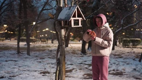 Young Woman Feeding Birds in Winter Park with Snowy Ground and Park Lights