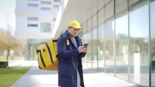 asiatischer männlicher Essenskurier benutzt Handy mit Handy-App auf dem Smartphone, während er in der Stadt auf der Straße steht