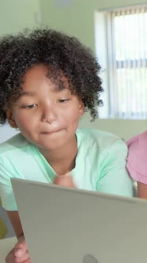 Vertical video: Leaning kid in pale green shirt responding to screen at classroom table with laptop