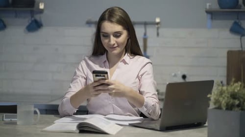 Happy young woman browsing internet on smartphone at home by table