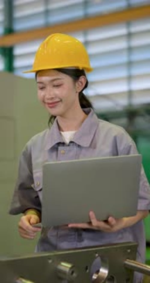 Asian adult woman engineer in yellow hard hat uses laptop beside metal mold, tracking production