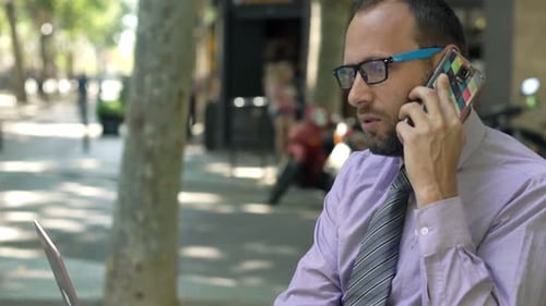 Angry Businessman with Laptop Talking on Cellphone Sitting on Bench in City 30s