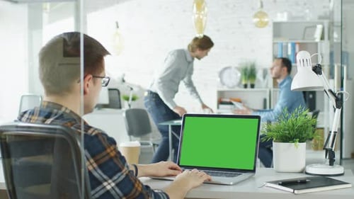 Close-up of a Man Sitting at His Desk with Green Screen Laptop. In Background Blurred and Brightly