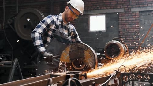 Male factory worker in hard hat and uniform is cutting metal by a big saw