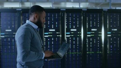 Black Data Center IT Technician Walking Through Server Rack Corridor with a Laptop Computer. He is
