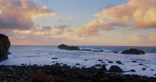Ocean waves crash on rocky seashore, sunset is spilling a golden tint into the fluffy clouds of sky