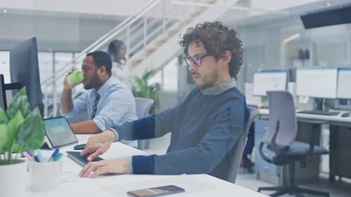 Young Handsome Manager with Curly Hair Works on a Desktop Computer and Compares Data with a Softwar