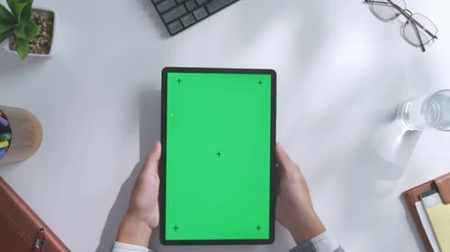 Top down view of hands holding a mock up green screen tablet on a white desk