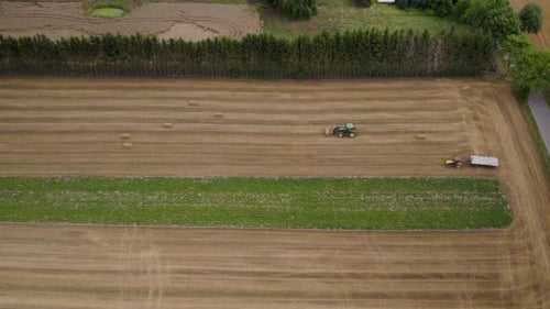 Industrial tractor picking hay bales from agricultural field and driving backwards - aerial top down