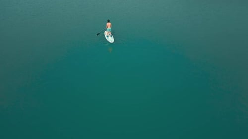 Aerial drone flying over a man doing stand up paddle in a blue water lake with mountains