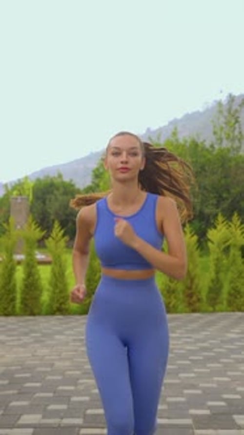 Young athletic woman in blue sportswear running outdoors on a paved path with trees and mountains