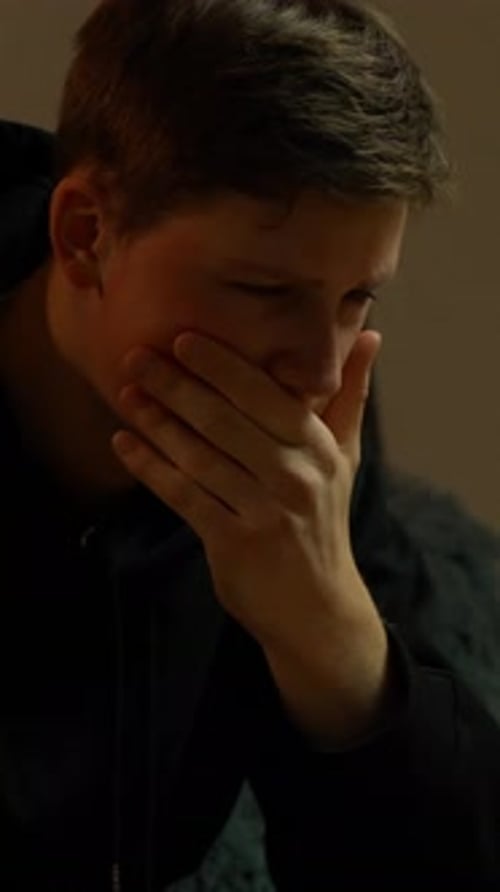 Young man covers his mouth in a dim room, showing deep emotional tension and inner struggle,
