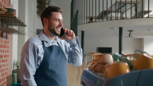 Caucasian Man Coffee Shop Employee in Apron Talk on Mobile Phone Behind Cafeteria Counter