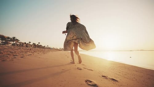 Sorglose, fröhliche Touristin in luftigem weißen Kleid, läuft bei Sonnenuntergang hinten am Meer entlang