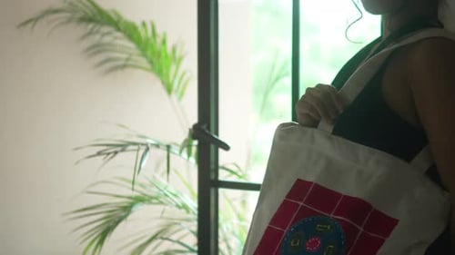 Mujer sosteniendo una bolsa reciclada, metiendo la mano dentro, de pie en una casa con una ventana y una planta