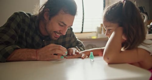 A Brunette Man in a Green Checkered Shirt Gives His Little Daughter a Brunette Girl in a Pink Dress