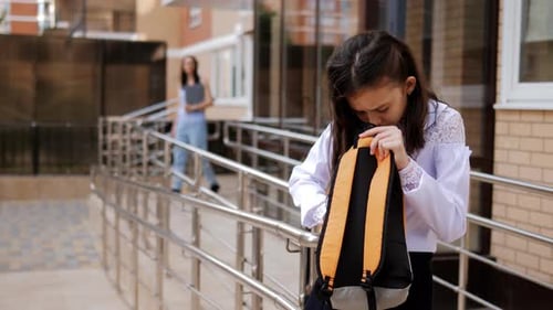 A Primary School Girl is Standing on the Porch of the School with a Backpack in Her Hands