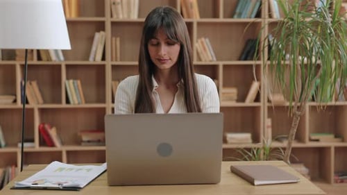 Woman Working at Laptop in Front of Bookshelf