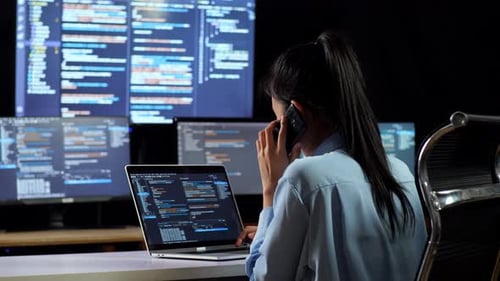 Visão traseira de uma programadora asiática falando no smartphone enquanto escreve código em um laptop