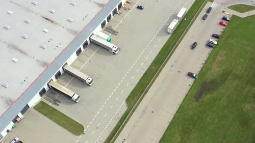 Aerial following shot of semi-trailer trucks travelling along a warehouse on the parking lot in the