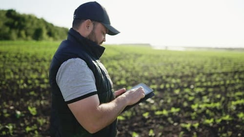 Farmer Stands and Smile Holds Tablet in His Hands Against Background of Working Tractor in Field