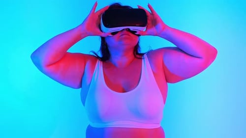 Woman with overweight in a tracksuit and VR headset posing in a studio with blue background and red