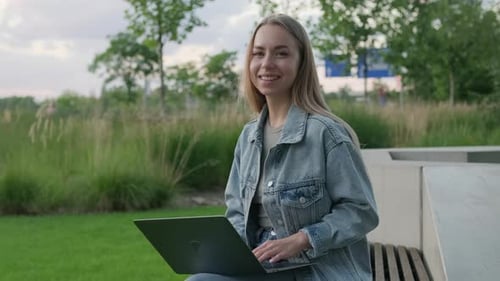 Young Woman Freelancer with Laptop on Lap Sits on Park Bench