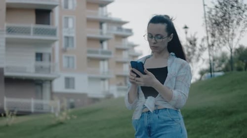 Young Woman with Eyeglasses Standing Alone in Residential Area Using Mobile Phone Looking at Screen