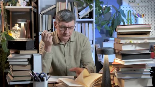 Mature Man Studying and Reading Books at Desk in Home Library