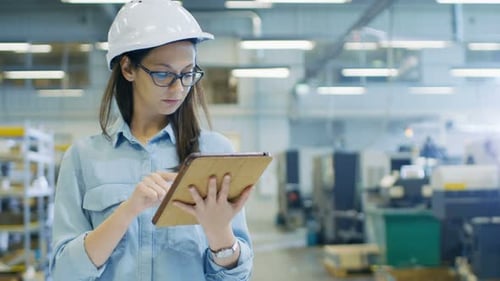 Female Industrial Engineer in a Hard Hat Uses Tablet Computer while Walking in Big Factory.