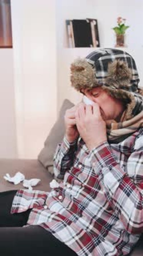 An Elderly Grayhaired Man in a Warm Earflap Hat Sneezes and Coughs Into a Napkin While Having the