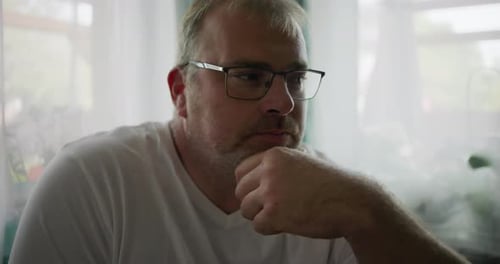 Man gazing pensively while resting his chin on his hand, seated near a window with soft green