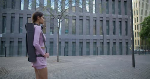 Eine schlanke, aktive junge Frau geht durch die Stadt und macht Pilates-Übungen mit einer Fitnessmatte, die an modernen...