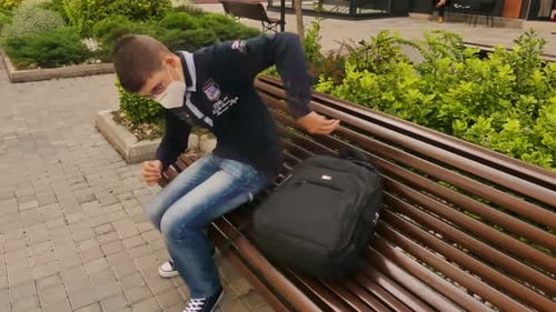 Teenage boy wearing face mask approaches to the bench, sit down and play on the mobile phone. Boy we
