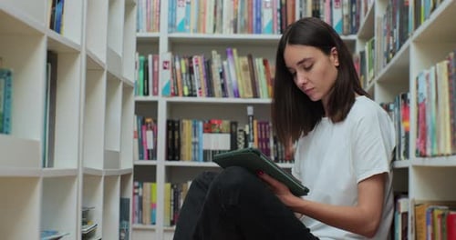 Caucasian Girl Sitting on the Floor Near Shelves with Book in the Library Uses Digital Tablet
