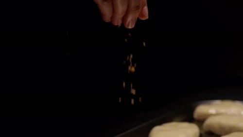Closeup of Human Hands Sprinkling Seasoning Over Raw Dough As Filled Buns Bake on a Dark Metal Tray