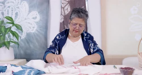 Una artesana asiática sénior mide la tela, cuenta las puntadas y luego dibuja el hilo para darle forma