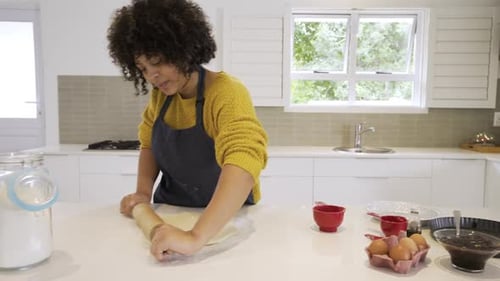 Woman wearing blue apron placing dough in kitchen, rolling it with rolling pin demonstrating baking