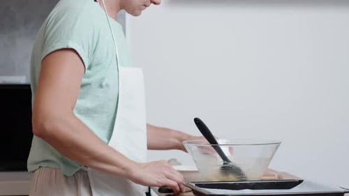 A woman in a white apron is preparing choux pastry. She cracks an egg into a bowl, viewed from the s