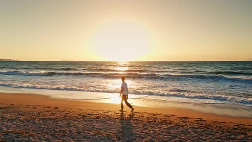 Young man with white shirt walking alone along the beach enjoying the beautiful sunset. waves coming
