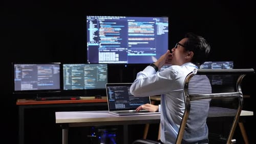 Back View Of Programmer Yawning And Sleeping While Writing Code By A Laptop Using Multiple Monitors