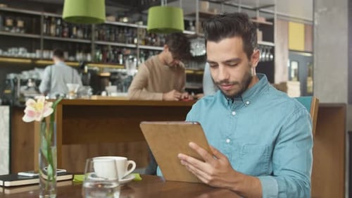 Hispanic Ethnicity Young Man using Tablet Computer at Cozy Coffee Shop.