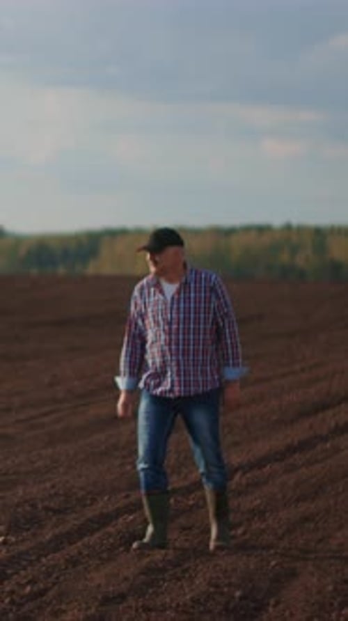 Solitary Farmer Walking Across Freshly Plowed Rural Field in Rubber Boots Inspecting Soil Under Open