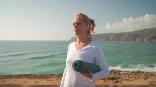 Happy Mature Woman with Rolled Yoga Mat Smiling at Beach