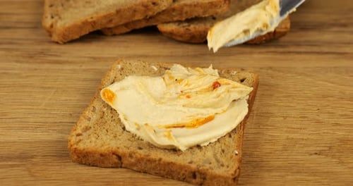 Le houmous est étalé au couteau sur un morceau de pain multigrains posé sur une planche à découper en bois.
