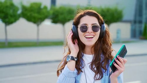 Portrait of cheerful young woman using headphones and modern smartphone for listening music